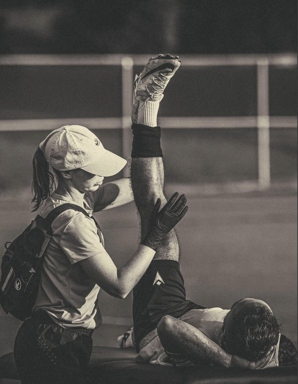 Ari supporting an athlete's raised leg through a hamstring stretch on a field