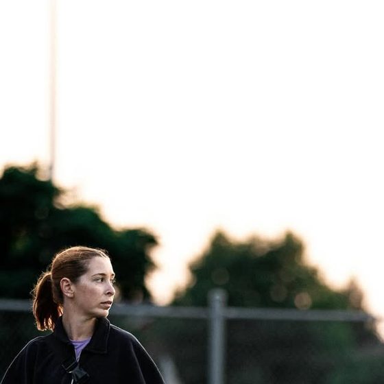 Ari outdoors in a jacket beside a sports field at dusk